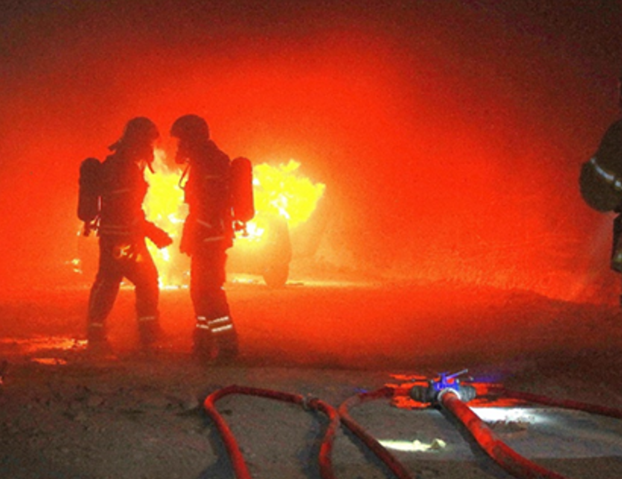 Zwei Männer in Feuerwehrausrüstung befinden sich untertage vor einem brennenden Fahrzeug.